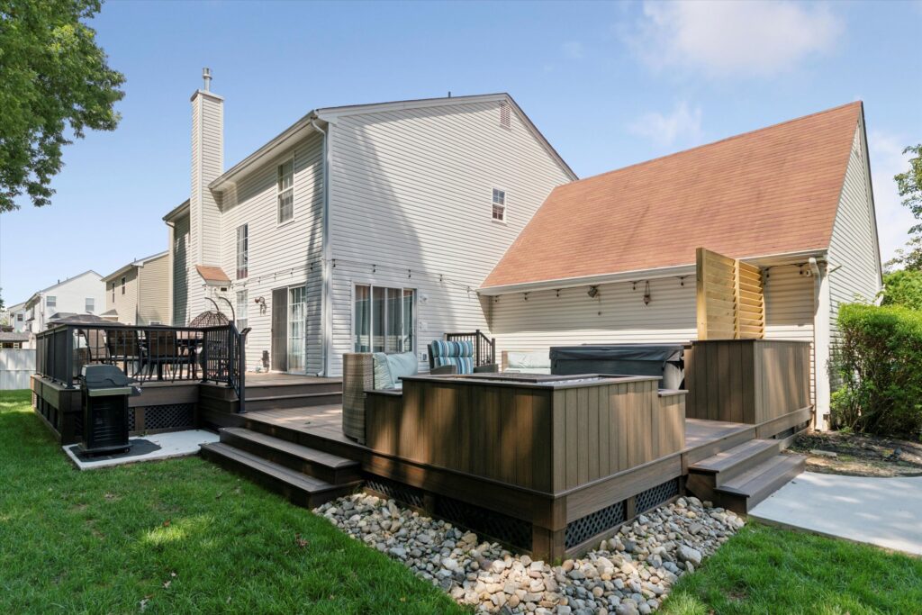 A custom-built, two-level deck in Burlington, NJ, constructed with Trex composite decking in a two-tone design using Toasted Sand and Spiced Rum. The deck features a built-in hot tub, outdoor seating areas, a new sliding door, and surrounding landscaping with a grass lawn and stone accents. in Burlington NJ 40.0705° N, 74.8662° W