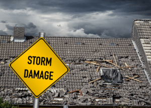 Severely storm-damaged roof with broken and missing tiles, scattered debris, and a dark cloudy sky in the background. A yellow road sign in the foreground reads 'STORM DAMAGE,' emphasizing the need for roof repair and restoration.
