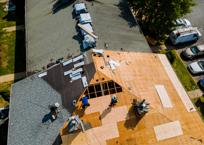 Aerial view of a roofing replacement project showing workers removing old shingles and installing new plywood sheathing. The roof is partially covered with roofing underlayment and new asphalt shingles, highlighting the roof repair and installation process.