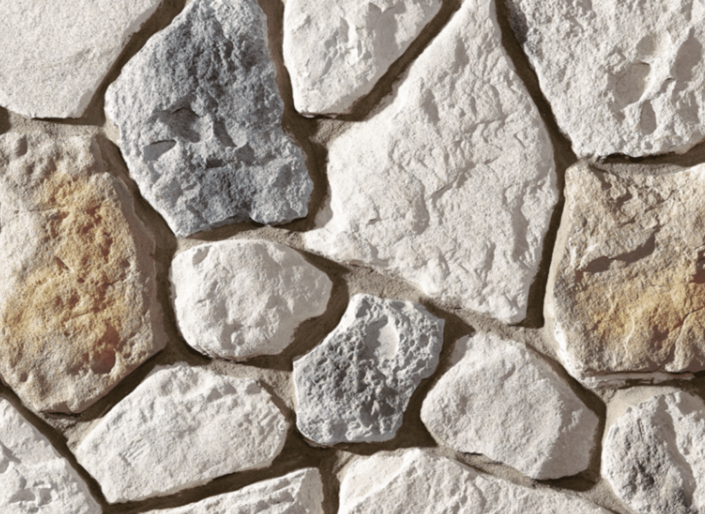 A close-up view of a fieldstone wall featuring irregularly shaped stones in shades of white, gray, and beige, arranged in an exterior stacked stone veneer style with visible mortar joints.