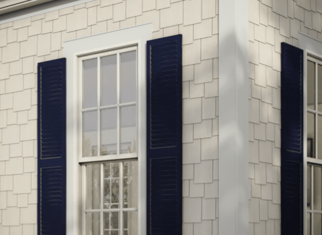 A close-up of a house exterior featuring Hardie® Plank Trim in crisp white, complementing light beige shingle-style fiber cement siding. The trim outlines large windows with deep blue shutters, adding a classic contrast. Sunlight casts soft shadows, highlighting the texture and durability of the materials.
