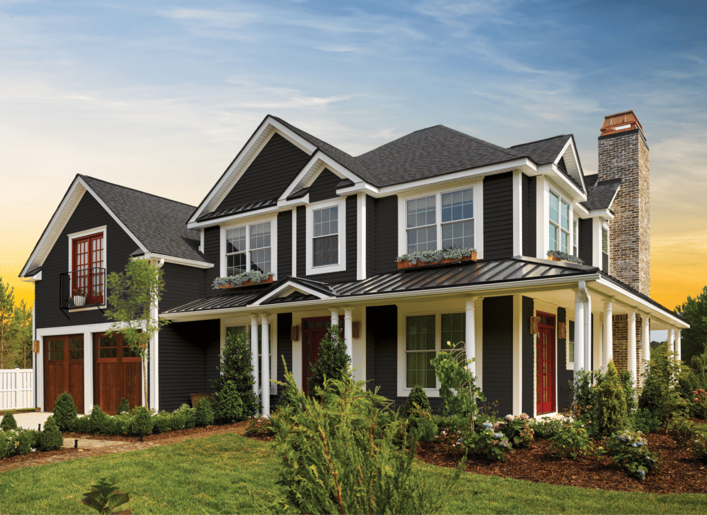 A beautifully remodeled home featuring newly installed dark CertainTeed siding with white trim, a wraparound porch, and a brick chimney. The well-maintained landscaping and warm sunset sky add to the inviting curb appeal.