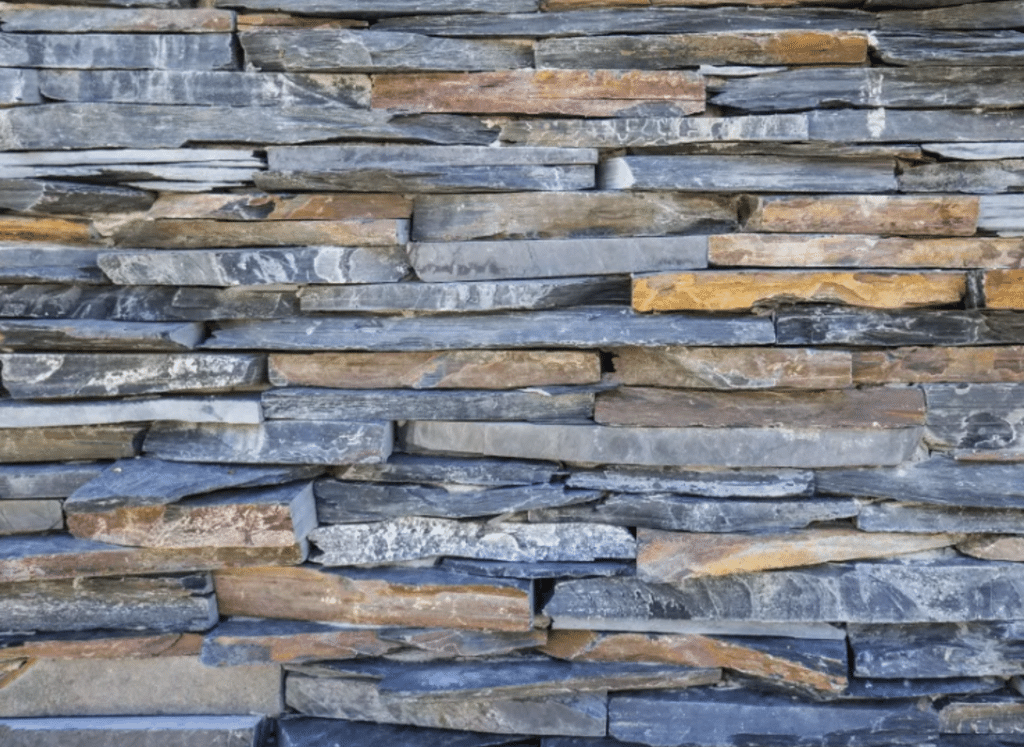 A close-up view of an exterior stacked stone wall made of slate stone veneer, featuring thin, layered stones in shades of dark gray, blue, and rust, arranged in a natural, textured pattern.