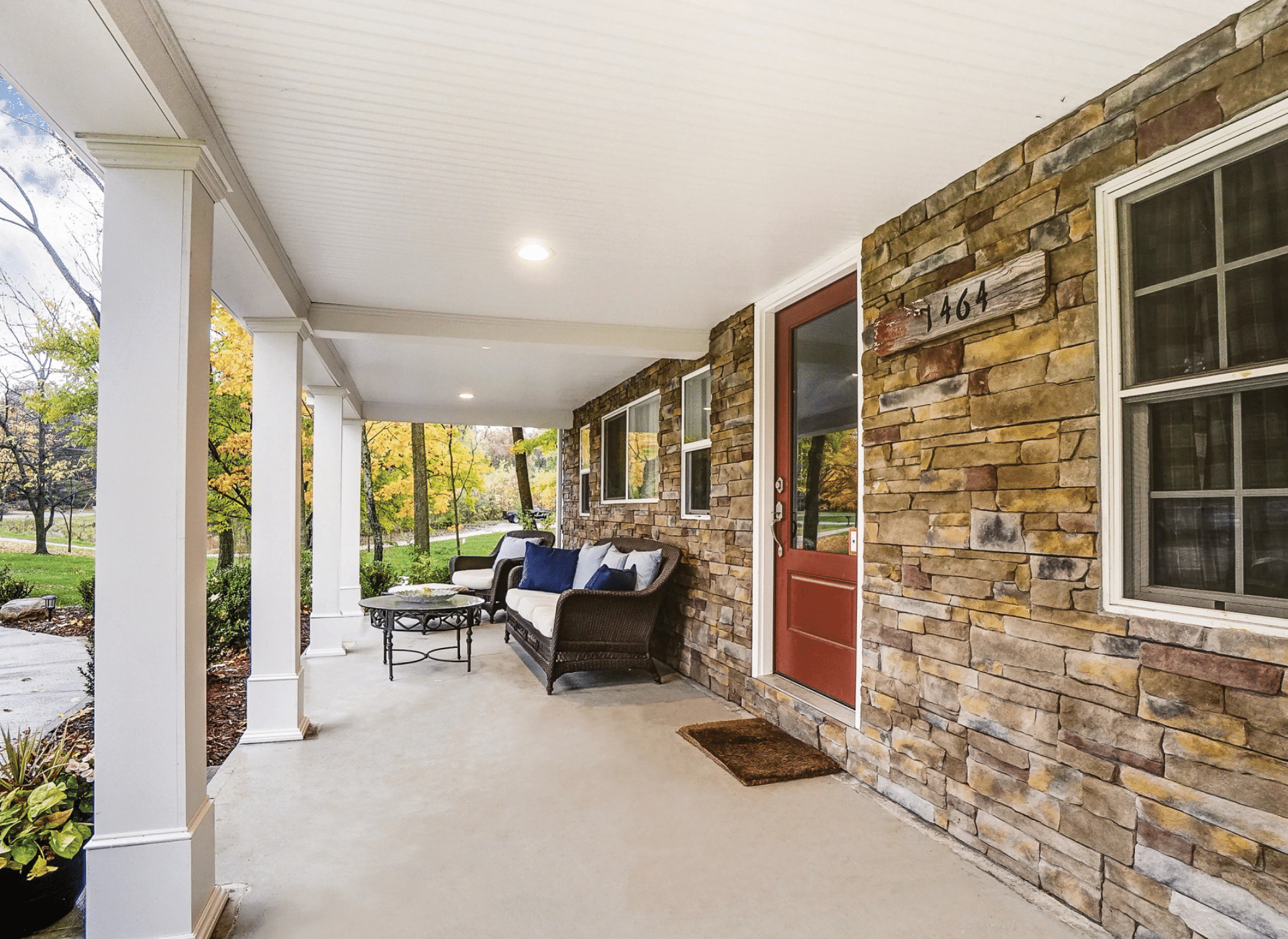 A cozy front porch with stone siding, a red door, and white columns. The porch is furnished with wicker seating, blue and white cushions, and a small table, overlooking a landscaped yard with trees showing autumn colors.
