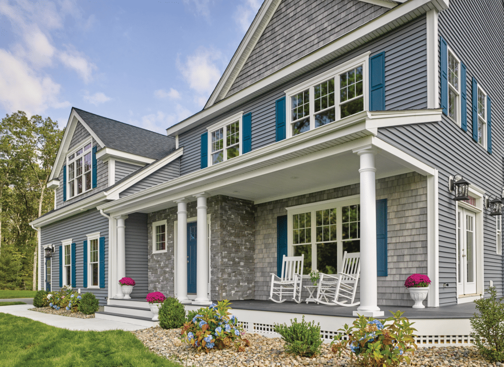 A beautifully designed home featuring CertainTeed vinyl siding in deep gray with white trim and blue shutters. The exterior combines horizontal siding, cedar shake accents, and stone veneer, with a welcoming front porch furnished with white rocking chairs and decorative columns.
