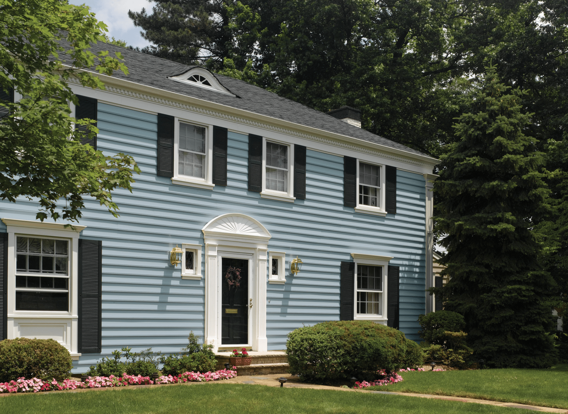 A classic colonial-style home with freshly installed light blue CertainTeed siding, white trim, and black shutters. The elegant entrance features a decorative white portico, flanked by gold lanterns, with a manicured lawn and blooming flowers.