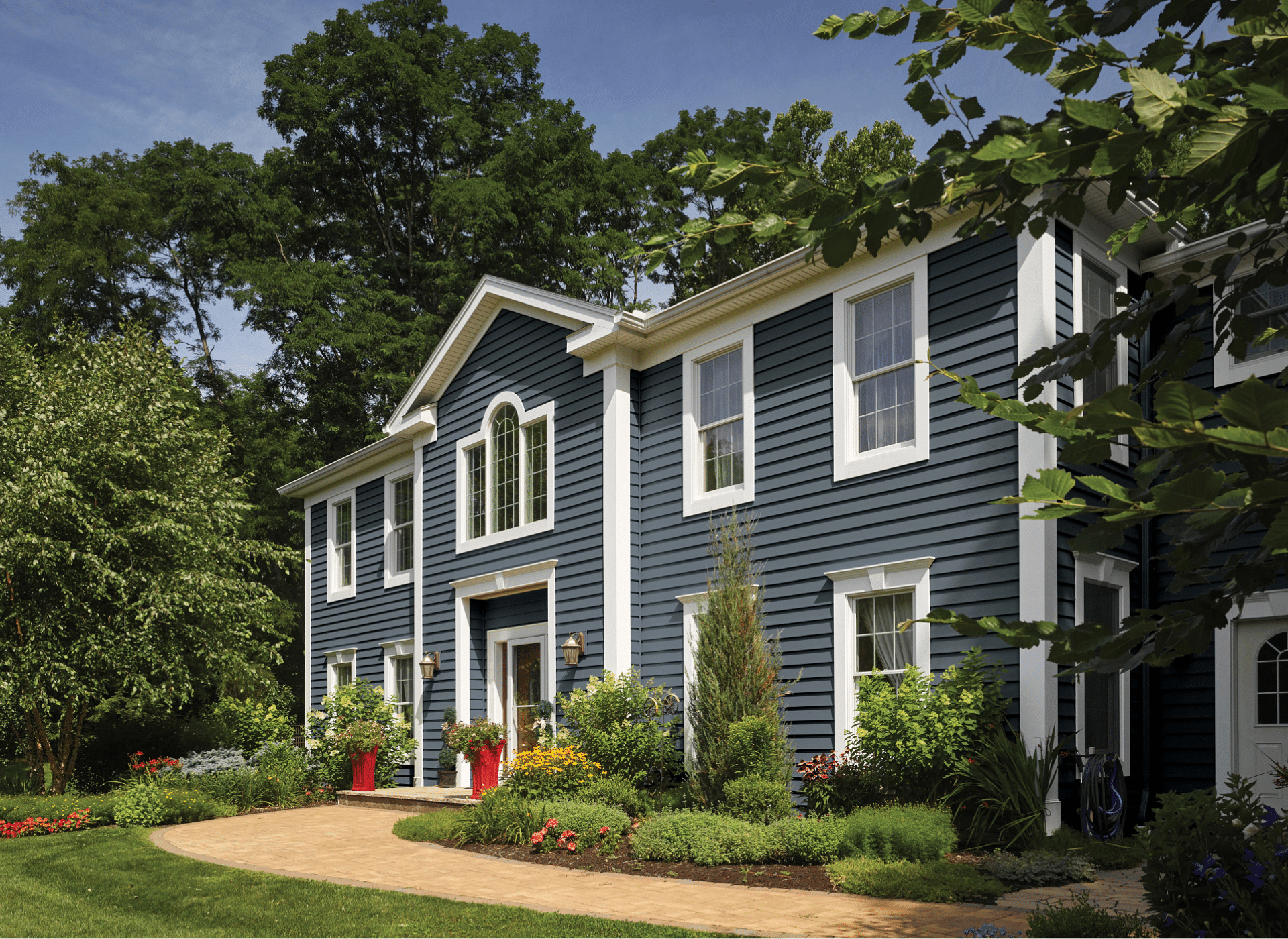 A beautifully maintained two-story home with deep blue CertainTeed vinyl siding, white trim, and an arched entryway. The exterior features a combination of horizontal siding and board and batten vinyl siding, complemented by lush landscaping and a curved walkway.