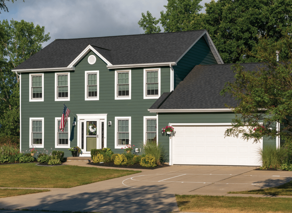 A colonial-style home with newly installed green CertainTeed CertaPlank siding, white trim, and a matching green front door. The house features a two-car garage, neatly landscaped yard, and an American flag displayed by the entrance.