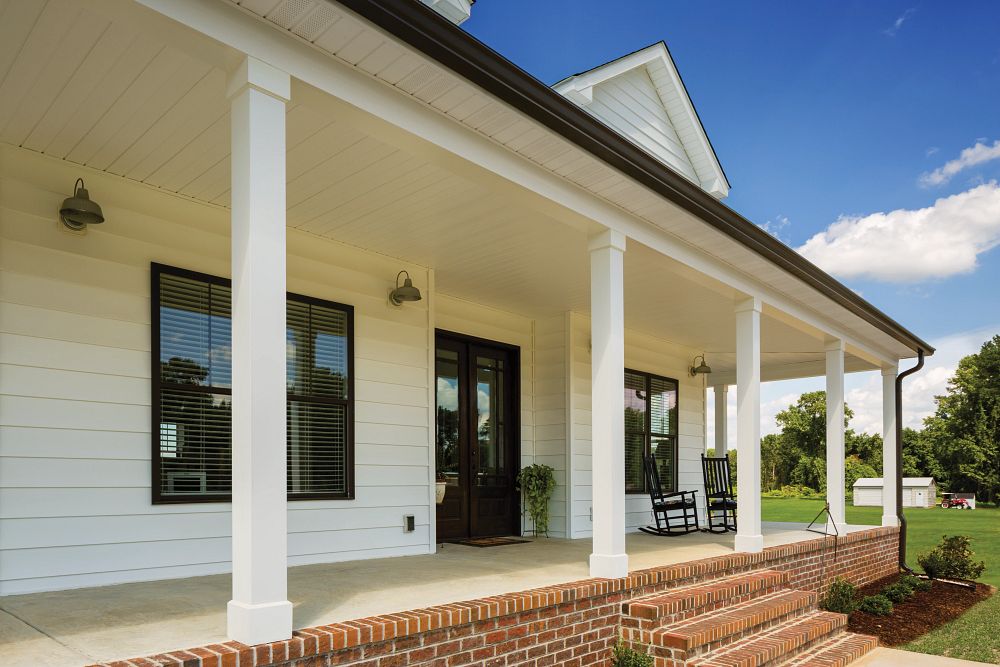 A house with fresh white vinyl siding replacement, featuring a spacious covered porch with white columns, black window frames, and a dark wooden front door. The porch has brick steps leading up to it, with black rocking chairs placed near the entrance. The home is surrounded by a well-maintained lawn under a bright blue sky.