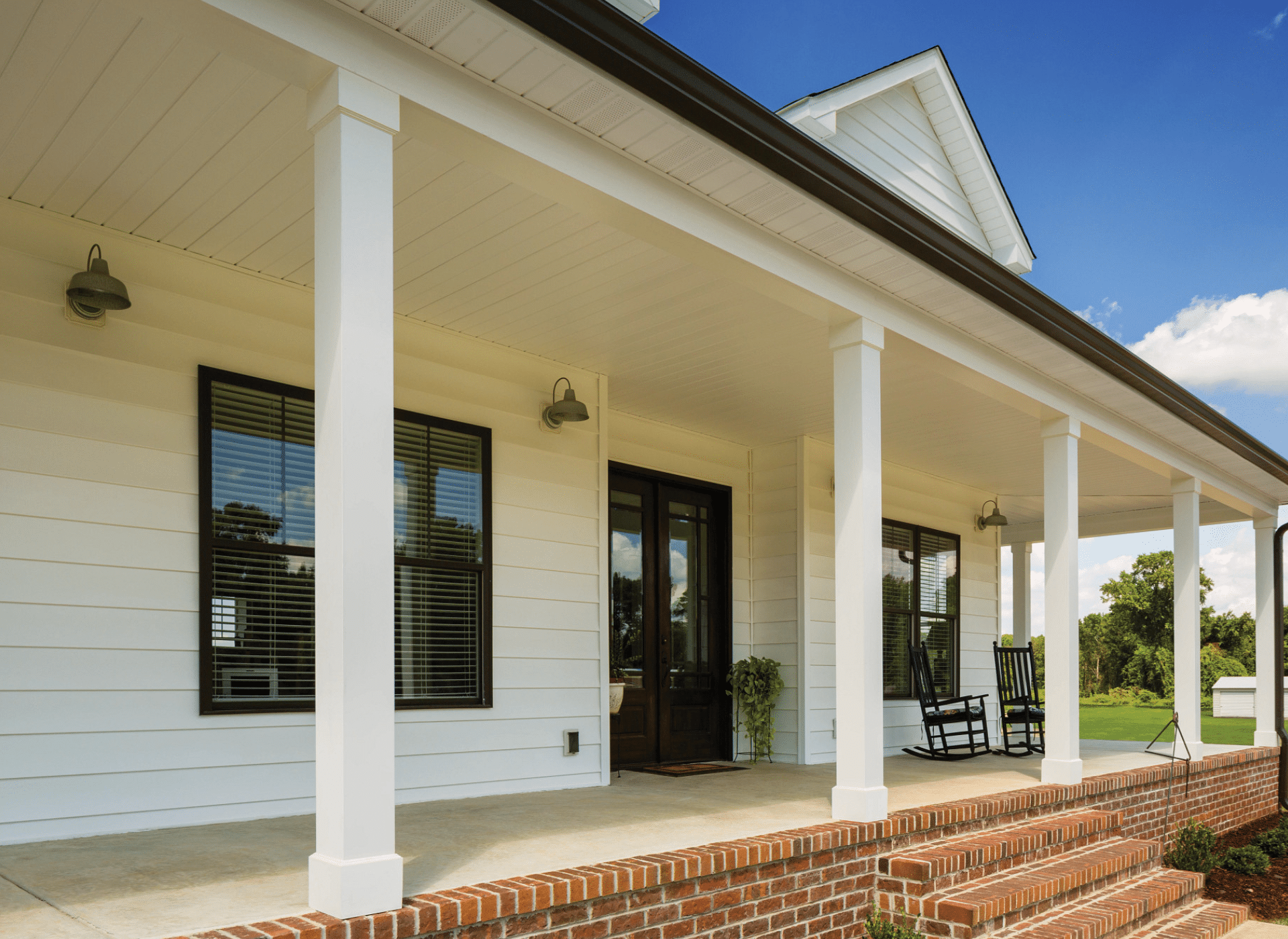 A charming front porch with freshly installed white vinyl siding, black-framed windows, and a dark wooden door. The covered porch features white columns, brick steps, and black rocking chairs, creating a classic farmhouse aesthetic under a bright blue sky.