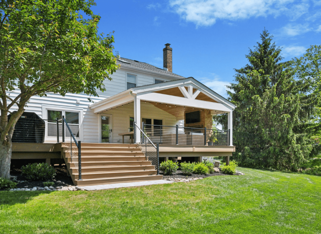 A luxious deck with a-frame roof covering on the rear of a large single-family home, a wide staircase and black aluminum cable railings.
