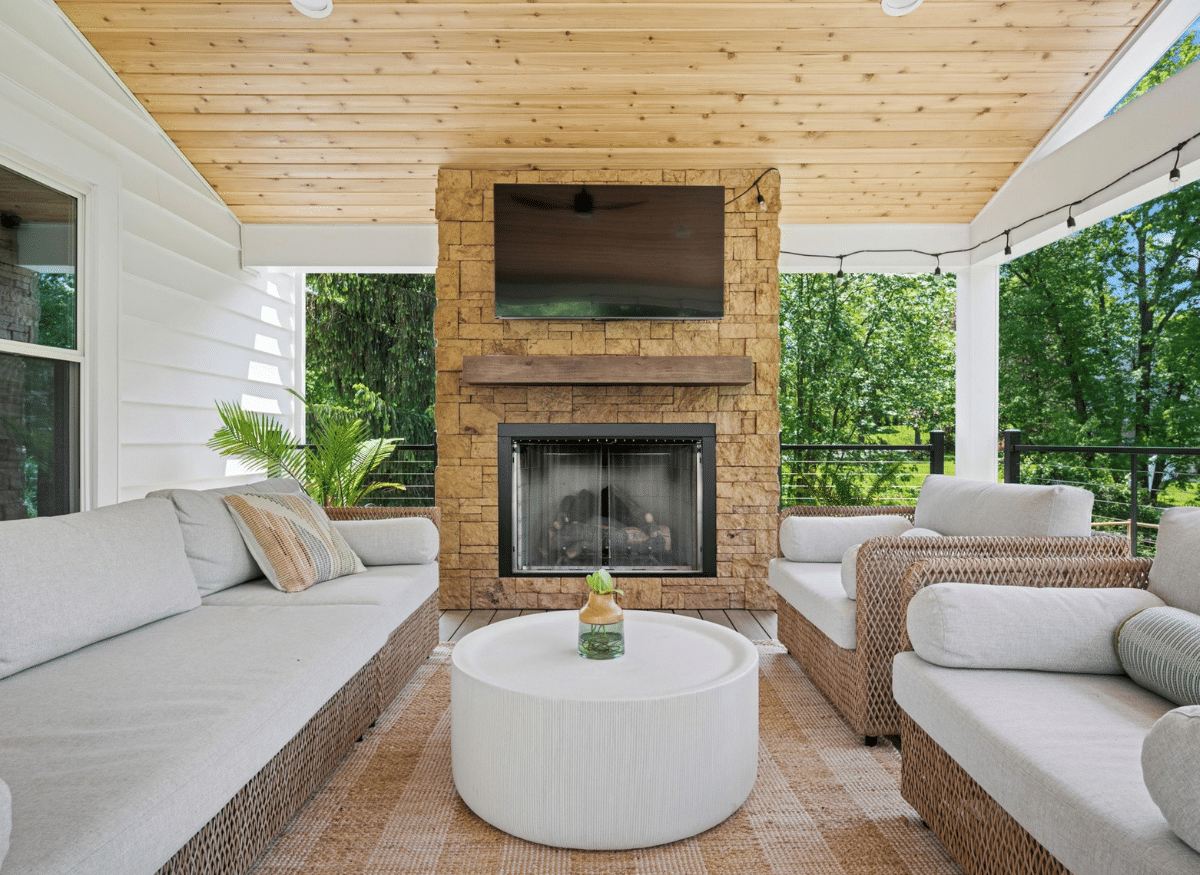 Cozy outdoor living space with wicker furniture, a stone fireplace, wood ceiling, and mounted TV under a covered deck surrounded by trees.